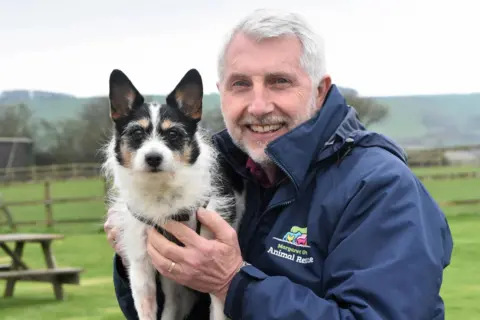 Margaret Green Animal Rescue Chris has short white hair and a grey/white beard. He is wearing a blue animal rescue centre jacket and is smiling and posing with a black and white dog.