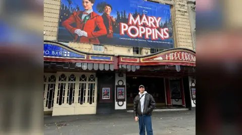 Rhys Batten Rhys Batten standing outside the front of the Bristol Hippodrome Theatre, with a poster for the show on the wall behind him