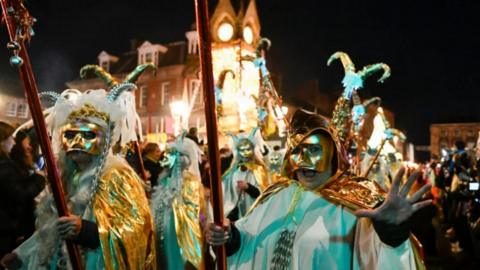 A night-time procession with people dressed up in colourful clothes and wearing masks.