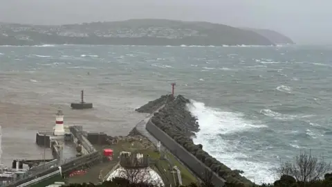 Grey skies over a choppy Douglas Bay, with waves crashing on the breakwater and Onchan Head in the distance.