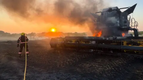 A firefighter in a field holding a hose with the sun setting, along with a burnt field and a combine harvester on fire. Trees are in the distance. Smoke is billowing up from the machinery. 