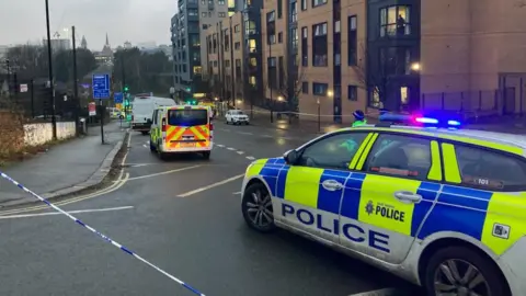 BBC A police car and police van on a road next to multiple apartment buildings. The road has been cordoned off with police tape.