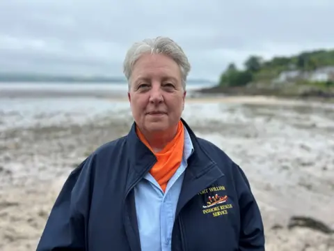 A woman with short grey hair standing on a sandy/muddy beach. She is wearing a blue jacket with a motif which says "inshore rescue service".