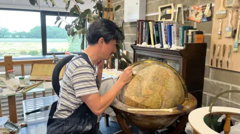 Tony Fisher/BBC A man in a workshop leans closely over a large globe, carefully applying detail with a small tool or brush. The globe, mounted in a wooden frame, shows an antique-style map with warm earth tones and fine lines. Around the workspace are paints, brushes, and a palette, along with shelves of books and neatly arranged tools on a wall. A large window behind the work area lets in daylight and reveals a green rural landscape outside.