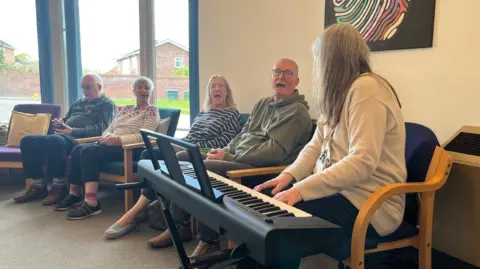 Music therapist Satyam is pictured playing an electric piano while a group of choir members sing.