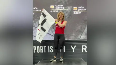 Contributed A woman stands on a podium. She holds a white flag out the side. She has short blonde hair and wears a red vest top with black leggings and running trainers. There is the number one spelled out on the front of the podium.