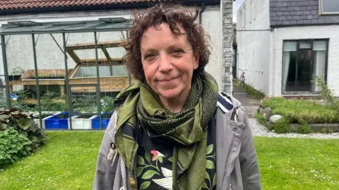 Caroline Williams is in a garden. She has dark hair and is wearing a green scarf over a grey jacket.