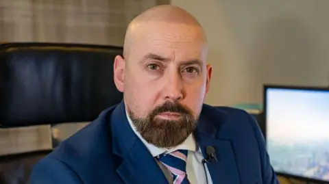 PA Media David Kennedy is bald with a brown beard. He wears a navy suit and sits in an office