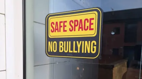 A shop window in Gateshead displaying a black vinyl sticker with a yellow and red font which reads "safe space no bullying". 