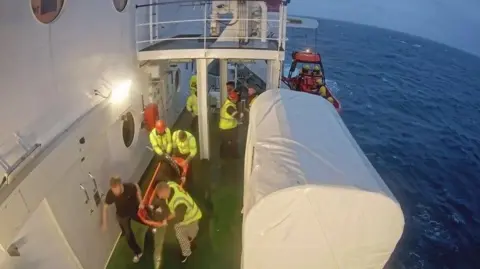 a screengrab for CCTV which shows a group of men in Hi-vis jackets carrying a man on a resuscitation board onto a ferry. The ferry is in the middle of a dark blue sea.