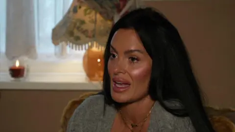 Keir Mackenzie/BBC A woman with black hair and wearing a grey shirt and gold necklace. She is sitting in a living room.