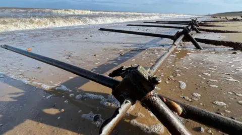 Dave Thurlow The remains of World War Two scaffolding poles emerging from sand at Minsmere in Suffolk. They formed the base of wartime defences. The metal poles are in a grid with a long section running parallel with the sea, while other poles are set at right angles and locked into position by joints. The sea is rolling back and there is a blue sky above. 