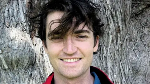 Ross Ulbricht, with hair blowing in the wind, pictured in front of a large tree trunk