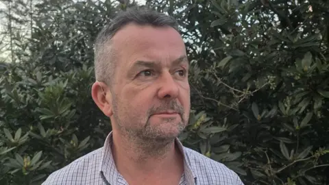 Glen Shaw is looking to the left and has grey and black stubble. He is wearing an open-necked shirt and there is a large, green bush behind him.