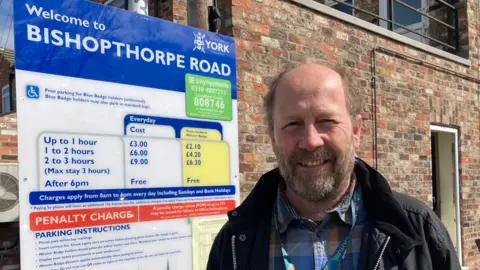 BBC/Jack Hadaway-Weller A prominent parking information sign is positioned on the left side of the frame, while a person stands to the right, close to the sign. The sign is predominantly blue and white, with additional sections in green, yellow, and red.