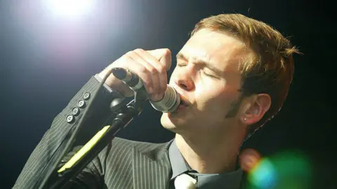 Getty Images Preston from the Ordinary Boys singing on a dark stage in close up. There is one bright white spotlight to the left. He is holding the mic in his right hand, close to his mouth, is wearing a grey suit and has his eyes closed