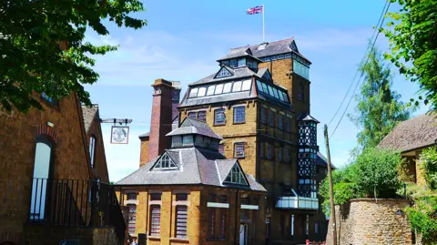 Hook Norton The exterior of Hook Norton brewery on a clear day. It is traditional Victoria tower brewery with brick walls and multiple floors.