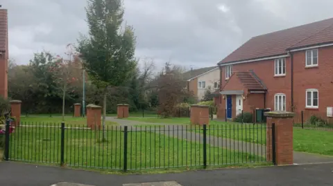 Carmelo Garcia An image of an open area in front of a red brick house, with the area enclosed by a black fence