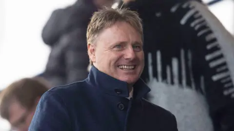 SNS Billy Hewitson, who has spiky blond hair, is wearing a dark jacket with the collar turned up. He is smiling.