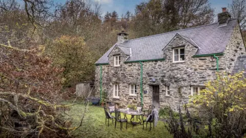 National Trust Mike Henton A two-storey stone farmhouse with a slate roof featuring two dormer windows. An outdoor dining table and chairs are visible in the garden in front of the house. Trees and shrubs surround the farmhouse and garden.