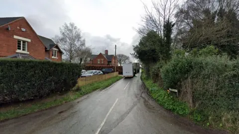 A generic image of a road, with bushes and trees to the left and right. There are buildings and vehicles parked to the left.