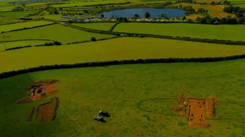 An aerial view of the excavation sites. Two sections of a green fields have been dug up, revealing the soil underneath.