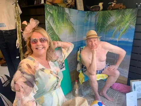 Alec W G Hall Ltd Debbie poses with her hand behind her hair and smiles. She wears a flowery dress with a fascinator hat. Andy is sat in a beach chair on a towel and wears a pair of swim shorts and a straw hat.