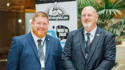 Supplied Image of Stu wearing a blue suit on the right. He is smiling and has ginger heard and a ginger beard. Alex is stood on the on the right with a bald head and grey goatee beard wearing a dark blue suit. They are stood in front of their charity branding with a plant in the background.