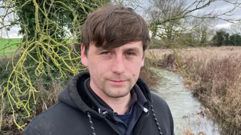 Connor Bryan, who has brown hair and is wearing a black hooded top, stands in front of a river.