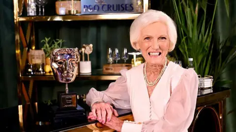 Mary Berry smiles at camera next to her Bafta trophy. She wears a pale pink dress, gold necklaces and has short grey hair. 