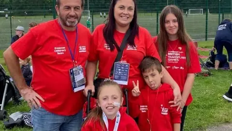 Young girl Elodie, who is sat in a wheelchair and has a tube going into her nose, poses with her parents and two siblings. The family are wearing matching red t-shirts and hoodies for the British Transplant Games.