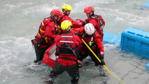 . Chris Jackson/Pool via REUTERS Five firefighters take part in a water rescue training scenario in the Northern Ireland Fire and Rescue Service’s (NIFRS) new Learning and Development College.  They men are standing knee-deep in water, dressed in red and black safety suits  and hard hats.  They are all holding an empty stretcher.