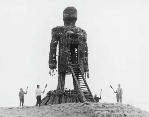 Getty Images The Wicker Man. A large wicker man standing in a field with four people standing beside it with torches, waiting to set it alight.