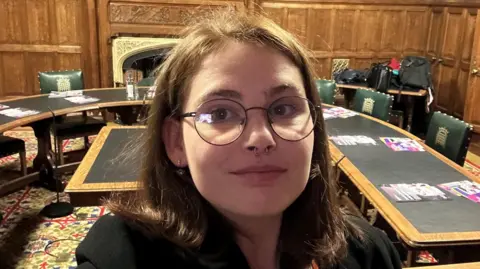 Stevie A young woman with brown hair and a septum piercing stands in a room in the House of Commons. Behind her are green leather chairs with gold crown emblems on them, and a long table.