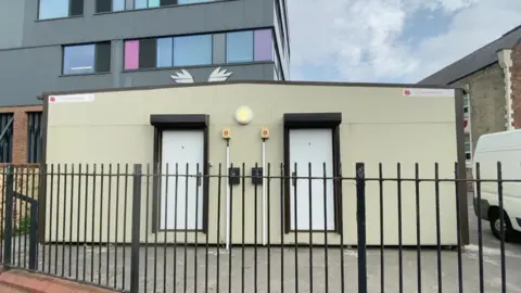 BBC/Richard Madden A cream modular building with two white doors in a car park behind black metal railings.