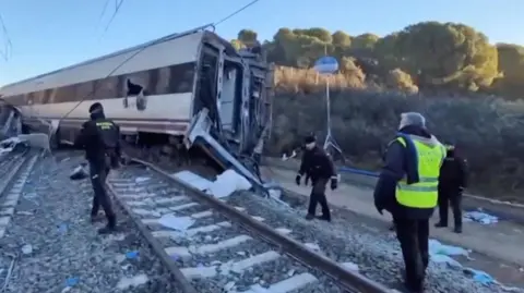 the scene of a train crash. A derailed carriage can be seen as police officers walk on the rails and embankment around it.