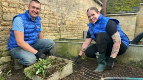 BBC Graeme and Katherine, both wearing blue gilets, kneeling down planting trees 