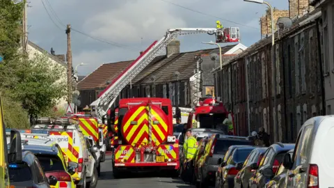 Wide image shows fire crews and emergency service vehicles parked on a terrace street dealing with the incident. 