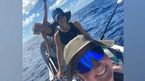 Row with the Flow/World's Toughest Row The picture shows three smiling people on a small ocean rowing boat in the middle of a vast stretch of deep blue water. One person is leaning out to the side of the boat with an arm raised, while another is seated and holding an oar. All are wearing sun hats. The sky is bright with scattered clouds, and the sea looks calm but expansive, giving a sense of isolation and adventure far from land.