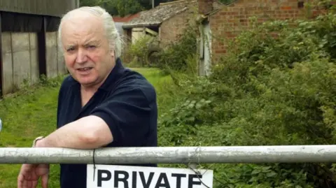 Tony Martin, pictured after his release, near a gate at Bleak House in Emneth Hungate, near Wisbech. He is wearing a black polo shirt and he has greying white hair. He has his left elbow resting on a gate, near which is a sign that reads PRIVATE. There are buildings in the background and grass and shrubs overgrown in the foreground.