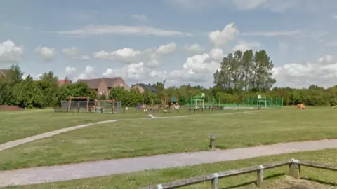 A large grass park with two grey footpaths and a large children's play park in the centre