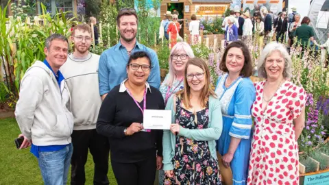J.Garget/University of Cambridge A group of eight people huddled in front of flowers, with two women at the front holding their award. They are standing together as part of a group and are all smiling at the camera in front of their exhibition.