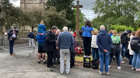 A crowd of people are stood around a road by a church.