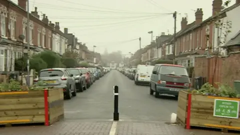 Street in Kings Heath, Birmingham, a bollard in the middle with two wooden boxes each side contains plants. Cars are parked on either side of the road next to houses.