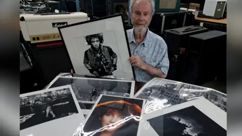 An older man with grey hair surrounded by glossy photos of pop stars, including Jimi Hendrix and Kate Bush. 