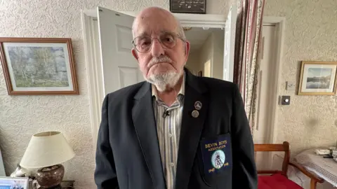 A bald man with a grey beard and moustache, frowning at the camera. He is wearing a white and blue pinstriped shirt and a navy blazer with a "Bevin Boys Association" logo. Behind him is a living room with a table, chair and lamp and pictures hanging on the walls.
