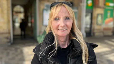 BBC/George Carden A blonde woman wearing a black puffer jacket and glasses resting on her head. She is standing outside a train station.