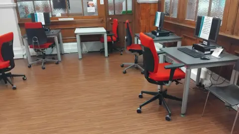 Contributed A room in an Edinburgh library with all computers switched off, with notices on the screen saying they cannot be used due to a 'technical fault'. The office chairs that are placed near the desks on the dark wooden floor are red, with a black backrest and frame.