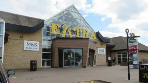 The entrance of the Extra services in Peterborough. A large brick building with a glass automatic door. Either side of the entrance are signs for M&S Simply Food and above the door is a large yellow sign that reads "ExTRA". 
