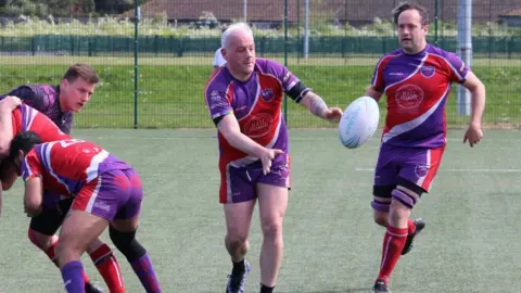 Northampton Outlaws A group of men playing rugby on an artificial pitch. There is a green wire fence behind them. Three men on the left are practising tackles and a man in the centre is being passed a rugby ball. They are all wearing red, white and purple kits.